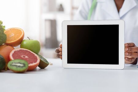Cropped of black doctor dietologist holding digital tablet with blank screen, fruits on tableの写真素材