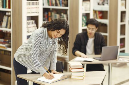 University Life. Mexican teacher writing in notebook, standing at desk in campus library, asian...