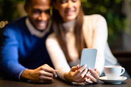 Technology And People Concept. Happy multiethnic couple sharing social media on smart phone sitting in coffee shopの写真素材