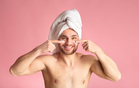 Male body care concept. Cute young man after a shower, white towel on head, smears cream on face.の写真素材