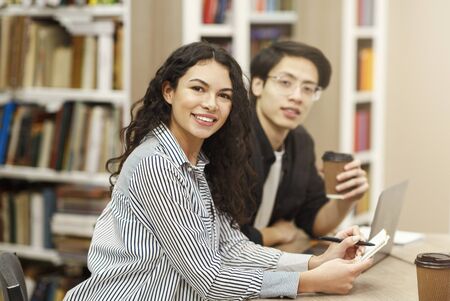 Student Life. Multicultural couple sitting at desk in university campus library, looking at camera, latin girl writingの写真素材