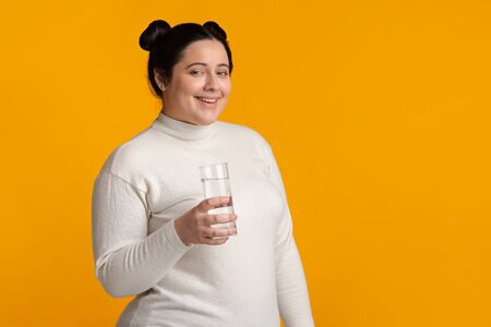 Hydration Concept. Young Positive Overweight Woman Posing With Glass Of Water In Hands Over Yellow BAckground, Free Spaceの写真素材