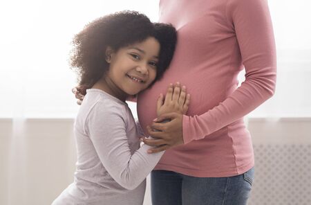 Adorable little afro girl embracing and touching pregnant mothers belly while standing together against window at homeの写真素材