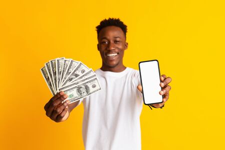 Excited african man holding lots of money in dollar currency and cell phone with blank screen in...