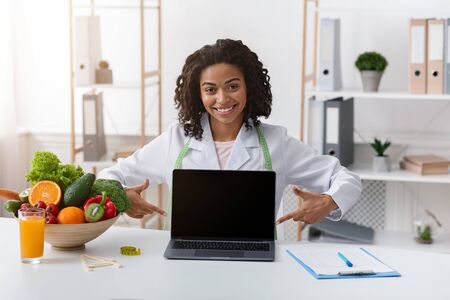 Cheerful afro woman dietologist pointing at empty laptop screen, sitting at her cabinet, nutritionist consultation onlineの写真素材