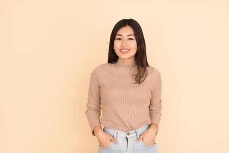 Cheerful asian girl smiling to camera and holding hands in pockets on peach background, free spaceの写真素材