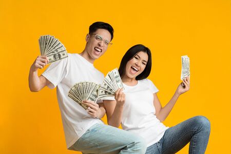 Super happy asian couple holding bunch of money over yellow studio backgroundの写真素材