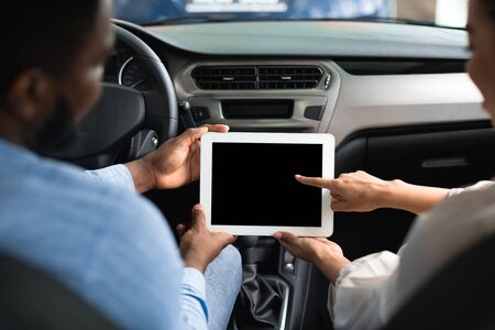 Afro Couple Using Digital Tablet With Empty Screen Sitting In New Car In Dealership Center. Mockup, Back View, Selective Focusの写真素材