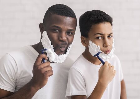 African American Father And Son Shaving In Bathroom Together, Holding Razors And Having Foam On Faces, White Background With Free Spaceの写真素材