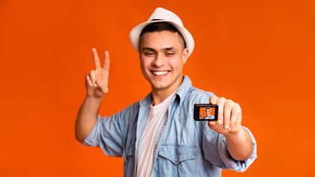 Happy handsome guy tourist posing and using action camera over orange studio background