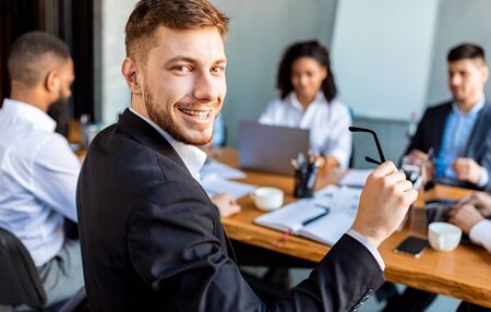 Business Career. Young Businessman At Corporate Meeting With Colleagues Smiling To Camera Sitting In...