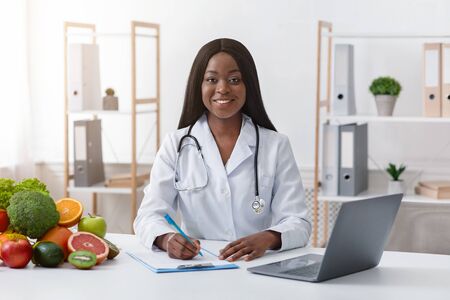 Smiling black woman dietologist working with laptop in modern office, having fresh fruits and vegetables on table, copy spaceの写真素材