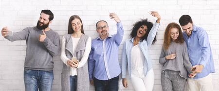 Social Media Addiction. Group of smiling people taking selfie with smartphones, posing over white brick wall, panoramaの写真素材