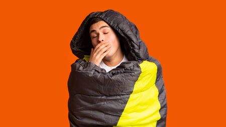 Sleepy guy tourist wrapped in sleeping bag yawning, orange backgroundの写真素材