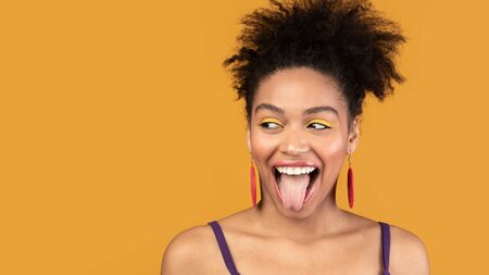 Having Fun. Young afro woman showing tongue and looking aside at copyspace isolated over orange...