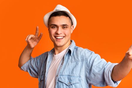 Positive guy traveler showing peace gesture and taking selfie over orange studio background