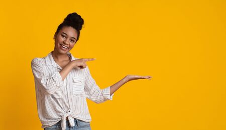 Cheerful Afro Woman Pointing At Something On Her Open Palm, Demonstrating Invisible Object, Free Place For Mockup, Yellow Background, Panoramaの写真素材