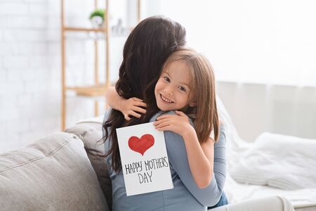 Smiling little daughter with Happy Mothers Day gift card hugging her mom, celebrating International Mothers Day together at homeの写真素材