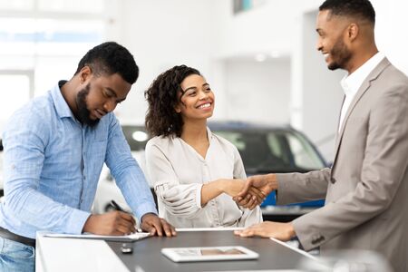 Buying Car. Black Couple Shaking Hands With Salesman And Signing Papers In Cars Selling Center. Selective Focusの写真素材