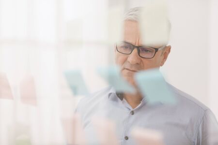 Business Strategy. Pensive Senior Manager In Specs Looking At Sticky Notes On Window At Office, Copyspaceの写真素材