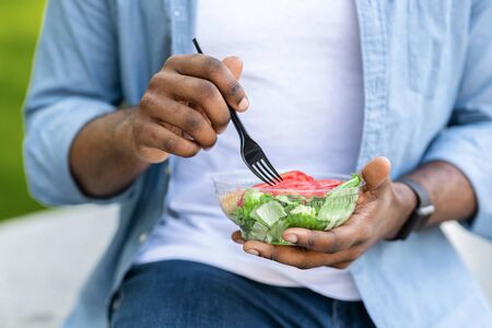 Healthy Lunch. Unrecognizable Black Man Eating Vegetable Salad Outdoors, Holding Plastic Bowl And...
