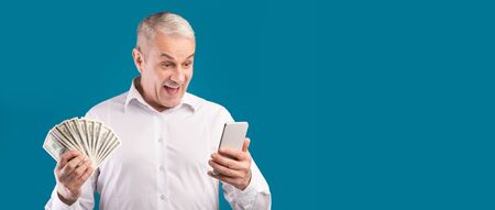 Make Money Online. Happy mature man holding smartphone and bunch of dollars, blue background,...
