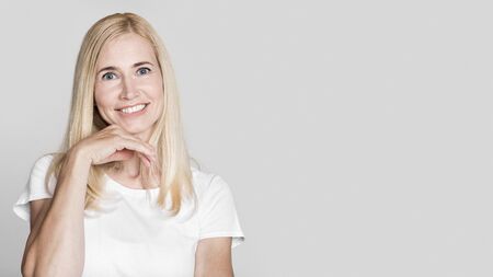 In A Good Mood. Smiling blonde middle-aged woman touching chin, looking at camera isolated over gray...