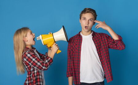 Siblings Quarrels. Annoying Little Sister Shouting At Her Elder Brother With Megaphone, Standing Over Blue Background In Studioの写真素材