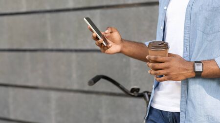 Unrecognizable afro guy with bicycle texting on smartphone and drinking takeaway coffee outdoors, panorama with copy space, cropped imageの写真素材