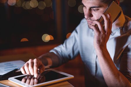 Business Guy Talking On Mobile Phone Using Tablet Computer Overnight Sitting At Workplace In Officeの写真素材