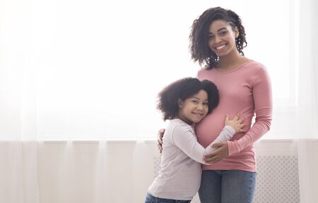 Happy expecting mom and her little afro daughter hugging next to window at home, looking at camera, copy spaceの写真素材