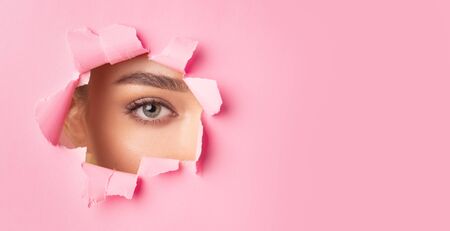 Unrecognizable Woman Looking At Camera Through Hole Torn In Pink Paper. Makeup And Beauty. Panorama, Empty Spaceの写真素材