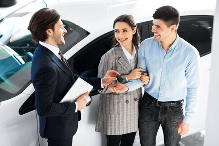 Automobile Purchasing Service. Couple Taking Key Of New Car From Cheerful Salesman In Auto...