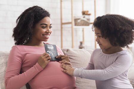Excited Pregnant Black Woman Showing Baby Ultrasound Scan To Her Little Daughter While Sitting Together On Sofa At Home, Closeupの写真素材