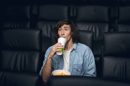 Cinematography concept. Millennial guy with drink and popcorn being captured by interesting movie in cinemaの写真素材