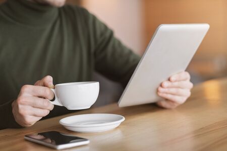 Close up of male hands holding cup of tea and digital tablet, working at cafe