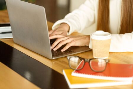 Girl works at laptop, cropped. Glasses, notebooks and coffee on the table
