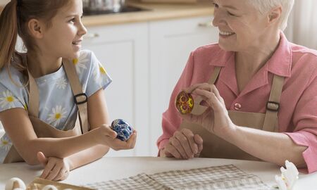 Happy holiday. Cheerful older lady and her adorable granddaughter with Easter eggs sitting at table...