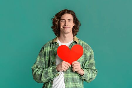 Expressing love. Romantic young guy holding red paper heart on color backgroundの写真素材