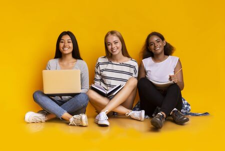 Three young cheerful multinational girls studying together with laptop, sitting on floor over yellow backgroundの写真素材