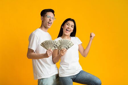 Young happy rich asian couple holding bunch of money over yellow studio backgroundの写真素材