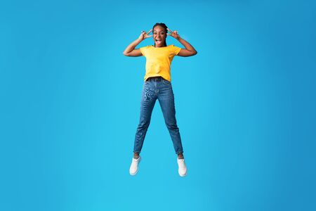 Joy Of Victory. Cheerful African American Girl Jumping Gesturing V-Sign Over Blue Studio Background....