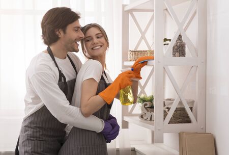 Domestic Duties. Young couple embracing while cleaning home together, wife and husband helping each...