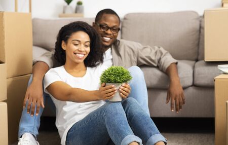 Moving to new place with many boxes. Smiling african american couple sitting on floor, girl holds...