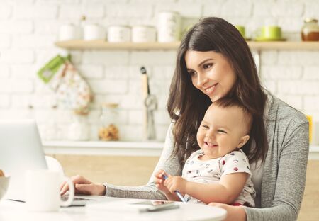 Maternity leave and business. Cheerful mother working on laptop at home kitchen, free spaceの写真素材