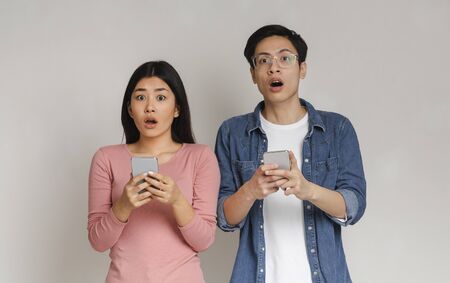Shocked asian man and woman holding smartphones over grey background, cheating onlineの写真素材