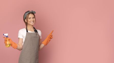 Cleaning Concept. Smiling young woman in apron holding detergent sprayer and pointing at copy space on pink background, panoramaの写真素材