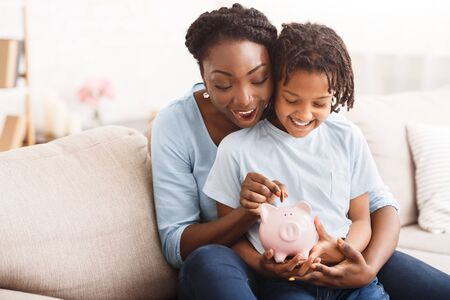 Money Saving Concept. Excited black mom and daughter putting coins into piggy bank, free spaceの写真素材