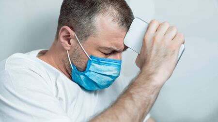 Self-Quarantine Depression. Desperate Man In Medical Mask Holding Phone Standing Over Gray Studio...