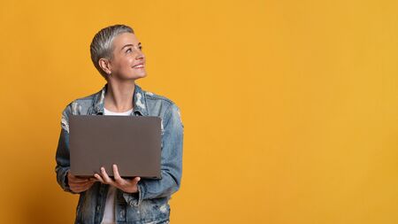 Smiling middle aged woman holding laptop and looking aside at copy space on yellow background,...